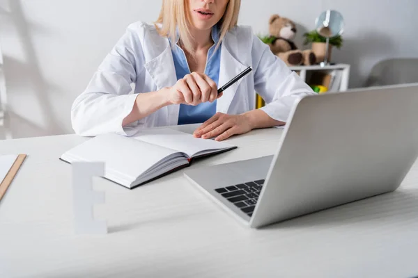 Vista recortada del terapeuta del habla en bata blanca que tiene videollamada en la computadora portátil en la sala de consulta - foto de stock