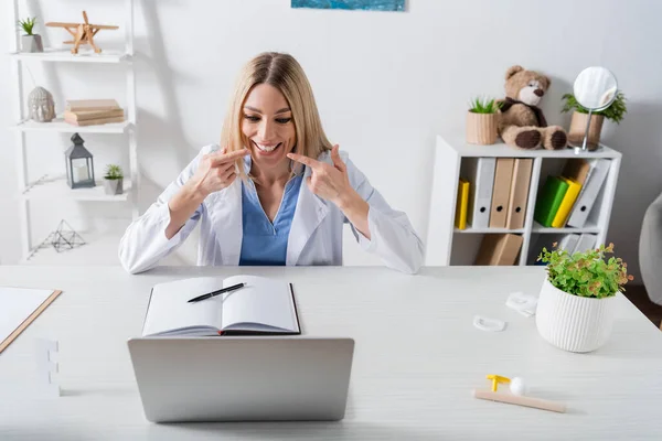 Speech therapist pointing at mouth during video call on laptop in consulting room — стоковое фото