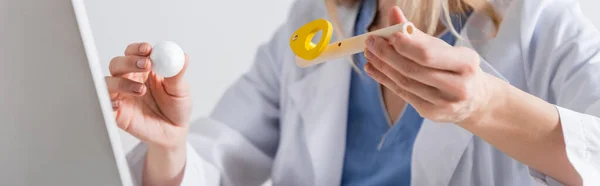 Vista recortada del terapeuta del habla en la capa blanca que sostiene el entrenador de músculo respiratorio cerca del ordenador portátil en la sala de consulta, pancarta - foto de stock