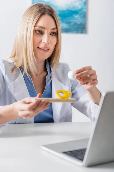 Logopedista positivo sosteniendo entrenador muscular respiratorio durante videollamada en portátil en sala de consulta - foto de stock