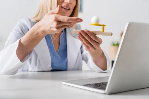 Vue recadrée du logopédiste pointant sur l'entraîneur de muscle respiratoire pendant l'appel vidéo sur ordinateur portable dans la salle de consultation — Photo de stock