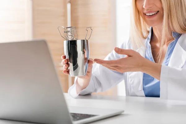 Ausgeschnittene Ansicht eines lächelnden Logopäden, der auf Werkzeuge in der Nähe eines verschwommenen Laptops im Sprechzimmer zeigt — Stockfoto