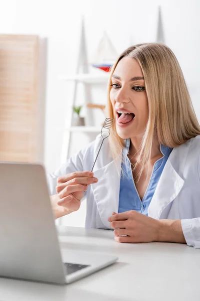 Speech therapist holding logopedic probe during video call on blurred laptop in consulting room — Stock Photo