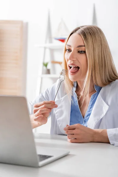 Orthophoniste tenant une sonde logopédique lors d'un appel vidéo sur un ordinateur portable flou dans une salle de consultation — Photo de stock