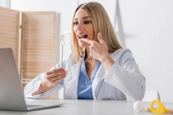 Speech therapist holding tool and pointing at mouth during video call on laptop — Stock Photo