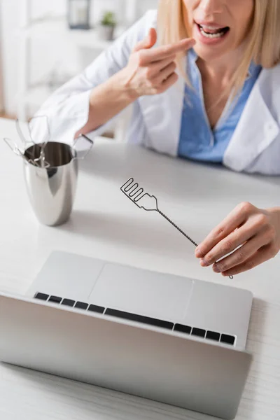 Cropped view of blurred speech therapist holding logopedic probe and pointing at mouth during video call on laptop — стоковое фото