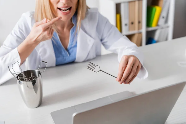 Cropped view of speech therapist pointing at mouth and holding tool during online lesson on laptop — стоковое фото