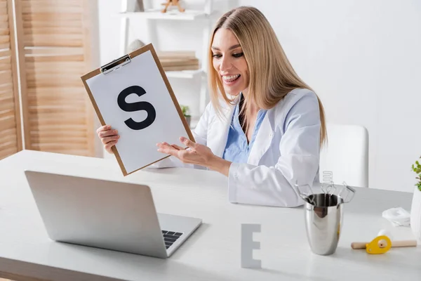 Positive speech therapist pointing at clipboard with letter during video call on laptop in consulting room — стоковое фото