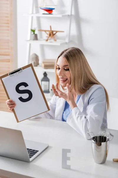 Speech therapist talking and holding clipboard with letter during video call on laptop in consulting room — Stock Photo