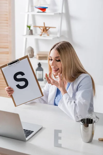 Speech therapist talking and holding clipboard with letter during video call on laptop in consulting room — стоковое фото