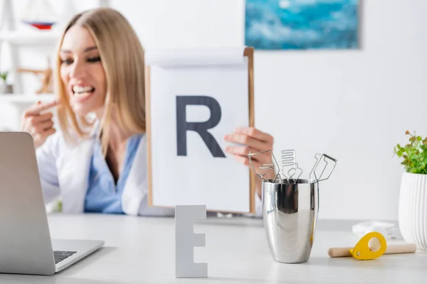 Carta y herramientas cerca de terapeuta del habla borrosa sujetando portapapeles cerca de la computadora portátil en la sala de consulta - foto de stock