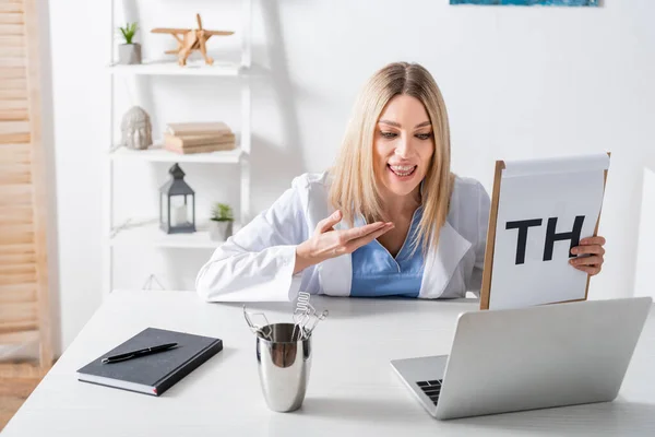 Logopedist pointing at clipboard with letters near tools and laptop in consulting room — Stock Photo