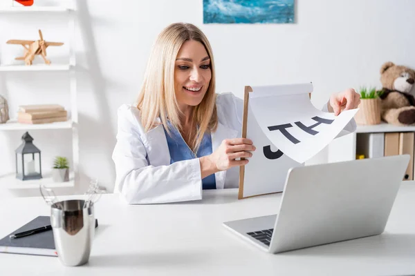 Joyeux logopédiste tenant presse-papiers avec des lettres pendant l'appel vidéo sur ordinateur portable dans la salle de consultation — Photo de stock