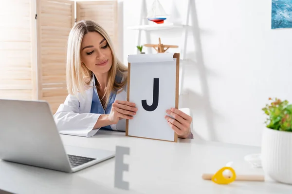 Speech therapist holding clipboard with letter during online lesson on laptop in consulting room — стоковое фото