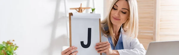 Logopädin hält Klemmbrett mit Brief neben verschwommenem Laptop im Sprechzimmer, Banner — Stockfoto