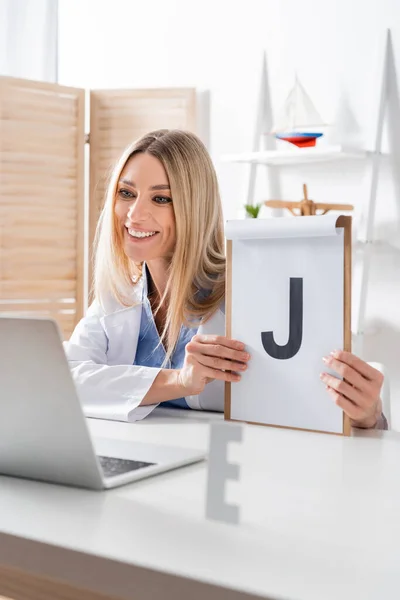 Happy logopedist holding clipboard during video call on laptop in consulting room — Stock Photo