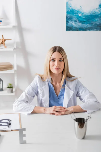 Terapeuta del habla mirando la cámara cerca de herramientas y portapapeles en la sala de consulta - foto de stock