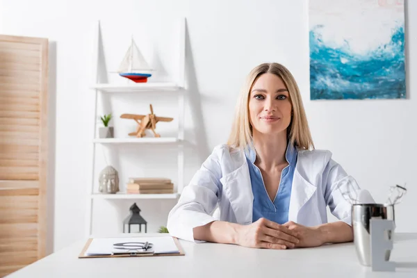 Logopedista en bata blanca mirando la cámara cerca del portapapeles y herramientas en la sala de consulta - foto de stock