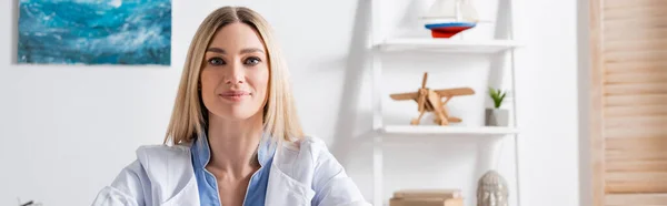 Logopedist in white coat looking at camera in consulting room, banner — Stock Photo