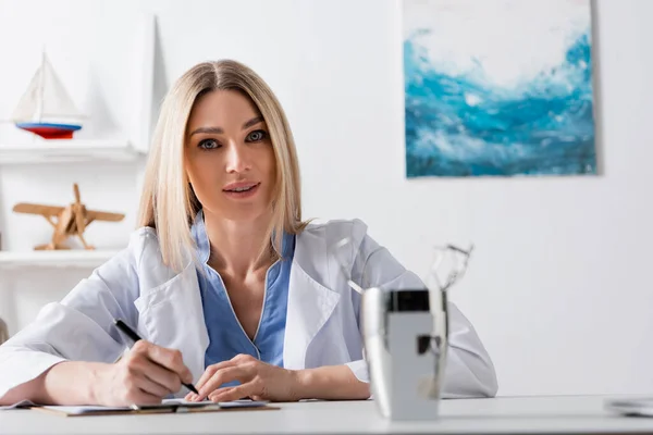 Speech therapist writing on clipboard near blurred tools in consulting room — Stock Photo