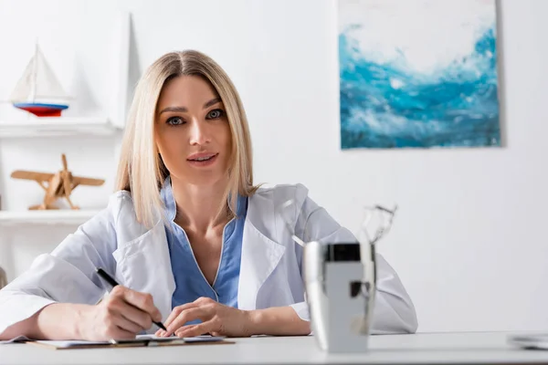 La escritura del fonoaudiólogo en portapapeles cerca de herramientas borrosas en la sala de consulta - foto de stock