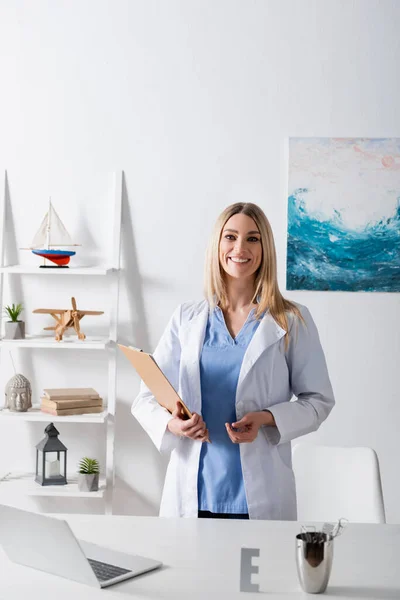 Cheerful speech therapist holding clipboard near laptop on table in consulting room — стоковое фото