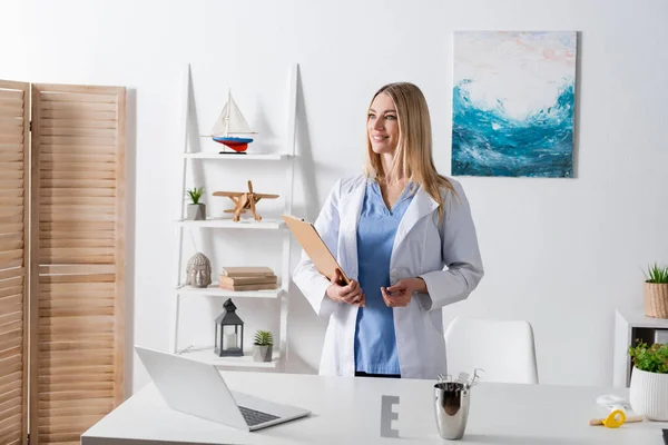 Logopedista alegre sujetando portapapeles cerca de herramientas y portátil en la sala de consulta - foto de stock