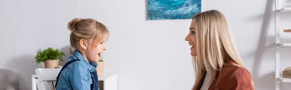 Vista lateral del niño hablando cerca de terapeuta del habla sonriente en la sala de consulta, pancarta — Stock Photo