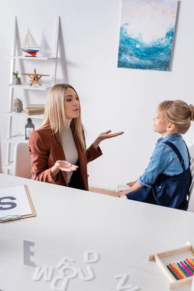 Terapeuta del habla hablando con el niño cerca de las letras y portapapeles en la mesa - foto de stock