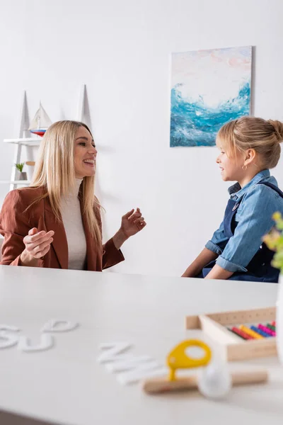 Logopedist talking with smiling girl near blurred letters and respiratory muscle trainer in consulting room — стоковое фото