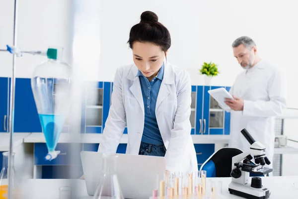 Joven genetista trabajando cerca de portátil y colega con tableta digital sobre fondo borroso - foto de stock