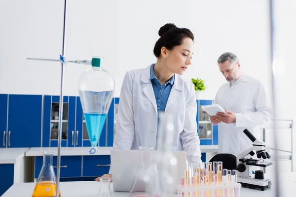 Bioengineer looking at microscope while standing near test tubes and flasks in lab — Stock Photo