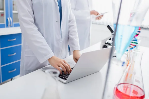 Vista recortada de científicos escribiendo en el ordenador portátil en el laboratorio cerca de colega borrosa - foto de stock