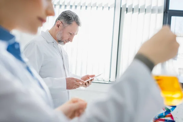 Beaded scientist using digital tablet near colleague on blurred foreground — Stock Photo