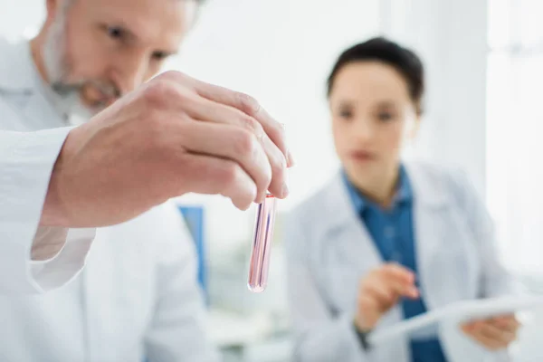 Bioingeniero sosteniendo tubo de ensayo con líquido mientras trabaja cerca de colega borroso - foto de stock