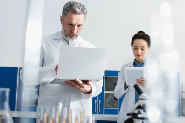 Bioengineers using laptop and digital tablet in lab on blurred foreground — Stock Photo