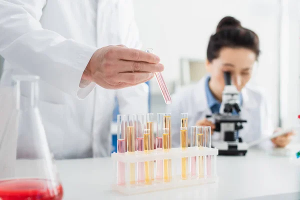 Geneticista segurando tubo de ensaio perto mulher borrada trabalhando com microscópio em laboratório — Fotografia de Stock