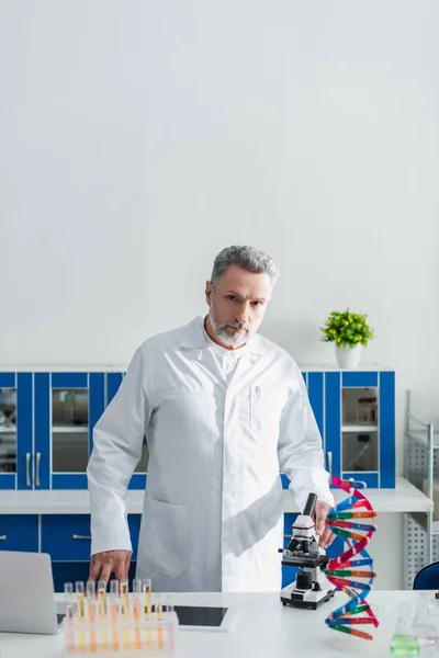 Généticien barbu en manteau blanc regardant la caméra près du modèle de l'ADN et du microscope — Photo de stock