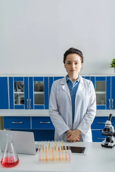 Joven bioingeniero mirando la cámara cerca de dispositivos y microscopio en el laboratorio - foto de stock