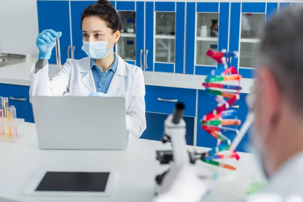 Geneticista em máscara médica trabalhando com tubo de ensaio e laptop perto colega turvo — Fotografia de Stock