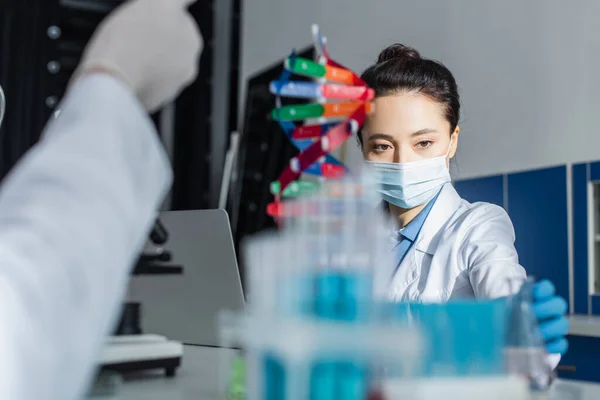 Genetista en máscara médica que trabaja cerca del modelo de ADN y colega borroso en el laboratorio - foto de stock