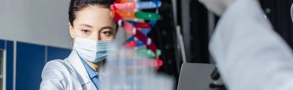 Генетик у медичній масці біля розмитої моделі ДНК в лабораторії, банер — стокове фото