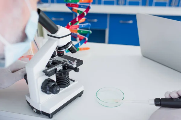 Partial view of blurred geneticist holding micropipette near petri dish while working with microscope — Stock Photo