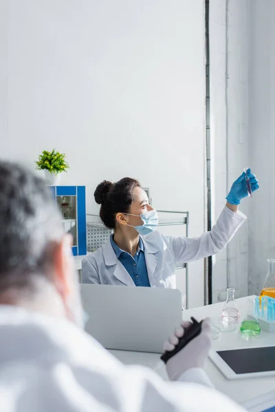 Biotecnólogo em máscara médica segurando tubo de teste perto de laptop e colega turvo — Fotografia de Stock