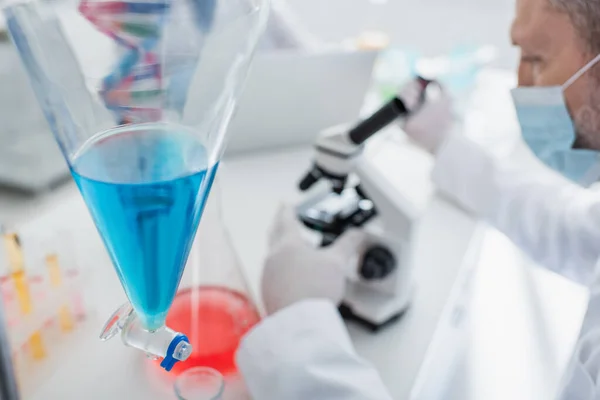 Foyer sélectif de la fiole avec du liquide bleu près du scientifique flou dans le masque médical — Photo de stock