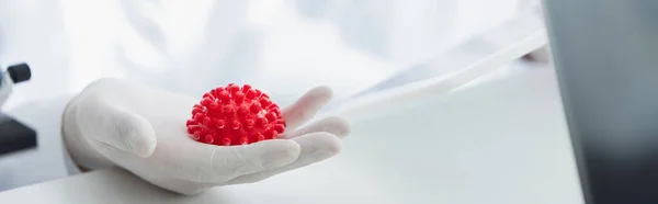 Vista recortada del virólogo en guante de látex sosteniendo el modelo de bacterias del coronavirus rojo, bandera - foto de stock