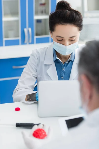 Geneticista em máscara médica trabalhando em laptop perto colega turvo e modelo de bactérias coronavírus — Fotografia de Stock