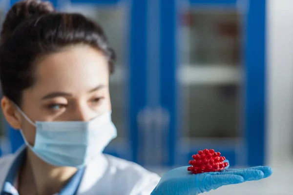 Blurred scientist in medical mask and blue latex glove holding red coronavirus bacteria model — Stock Photo