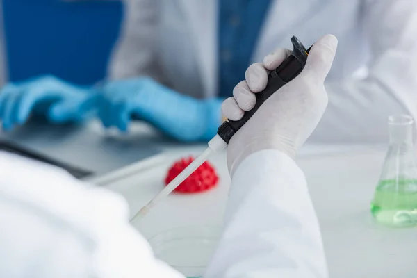 Vue recadrée du scientifique dans un gant en latex tenant une micropipette près d'un modèle flou de bactéries coronavirus — Photo de stock