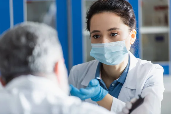 Bioingeniero en máscara médica y guante de látex apuntando con la mano mientras habla con un colega borroso - foto de stock